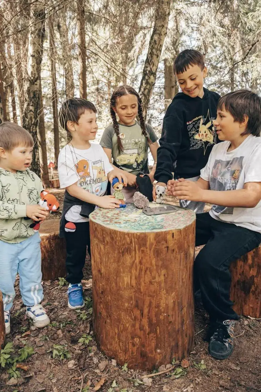 Pět dětí u dřevěného pařezu na pohádkové stezce v lese, hrají si s plyšovými skřítky